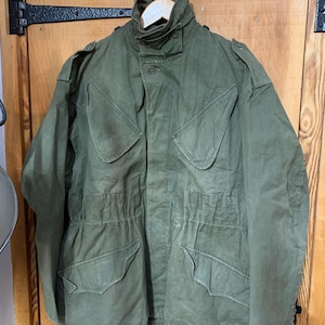 May include: An olive green military-style jacket with multiple pockets and a high collar. The jacket is hanging on a wooden hanger. The jacket has a button-up front and a drawstring waist. The jacket is made of a durable fabric.