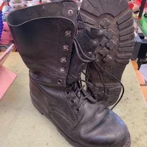 Puede incluir: Un par de botas de combate de cuero negro con diseño de caña alta, ojales metálicos, cordones negros y una suela de goma gruesa con tacos profundos para mayor tracción. Las botas tienen una puntera redondeada y soporte estructurado en el tobillo.