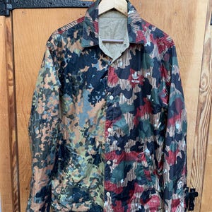 Puede incluir: Chaqueta Adidas reversible con estampado de camuflaje en tonos marrón, verde, rojo y negro. La chaqueta tiene cierre de botones, cuello y el logo de Adidas en el pecho. Cuelga de una percha de madera.