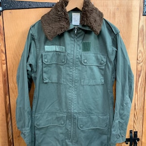 May include: A green military-style jacket with a brown faux fur collar. The jacket has multiple pockets and a zipper closure. The label on the jacket reads "98 L PM".