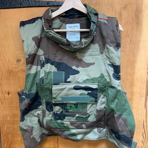 May include: Camouflage pattern vest with a high collar and multiple pockets. The vest is made of a durable fabric and features a zipper closure. The vest is hanging on a wooden hanger. The vest has the text "PAUL BOYE" and other text.