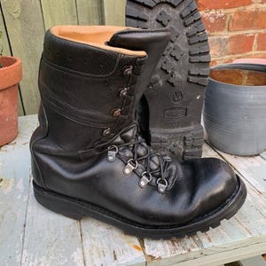 Puede incluir: Botas de combate de cuero negro con suela resistente. Las botas tienen ojales metálicos para los cordones y un cuello acolchado. Una bota está de pie, la otra muestra la suela.
