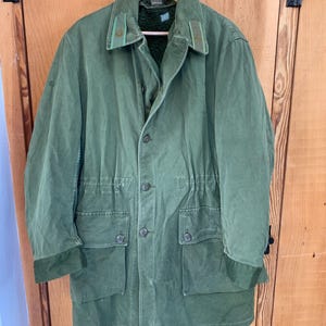 Puede incluir: Una chaqueta verde de estilo militar con cierre de cremallera y botones, dos bolsillos grandes y cintura con cordón. La chaqueta tiene un cuello de pana y un forro polar.