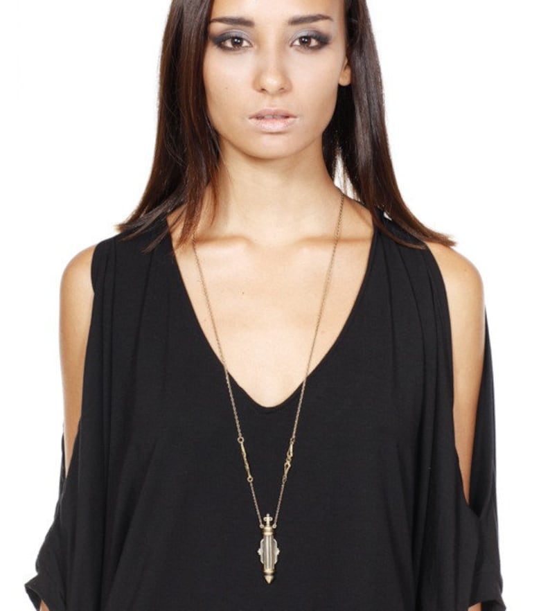 May include: A gold chain necklace with a decorative pendant shaped like a small, ornate bottle. The bottle is clear and has a black and white striped design.