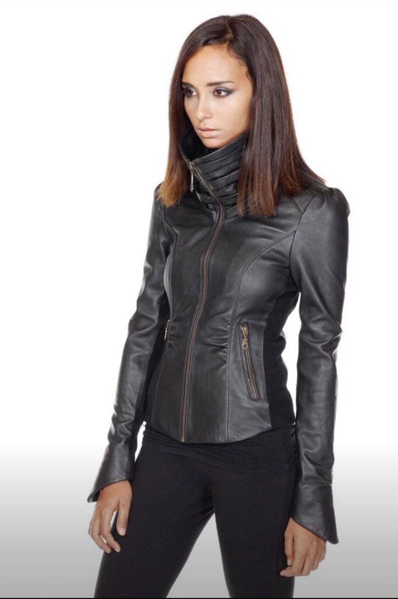 Puede incluir: Chaqueta de cuero negro con cuello alto y cierre de cremallera. La chaqueta tiene mangas largas y una silueta entallada. La modelo lleva pantalones negros. El fondo es blanco.