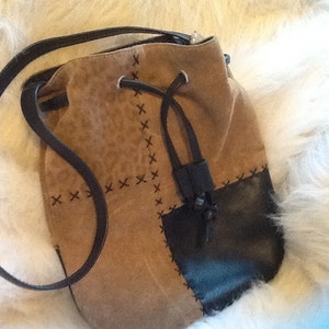 May include: A brown suede bucket bag with a black leather bottom and a drawstring closure. The bag has a long strap and is decorated with black stitching in an X pattern.