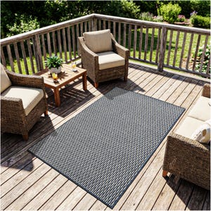 May include: An outdoor rug with a woven pattern in shades of gray and white, framed by a dark blue border. The rug is rectangular and placed on a wooden deck, with wicker chairs and a small table nearby. The scene suggests a patio or outdoor living space.