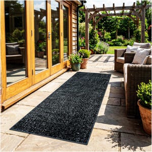 May include: A long, rectangular black outdoor rug with a textured surface. The rug is placed on a stone patio, near a wooden door and outdoor seating. The rug is designed for outdoor use.