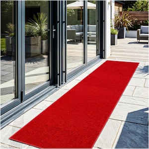 May include: A long, rectangular red outdoor runner rug. The rug is placed on a stone patio, leading towards a glass door. The rug is made of a textured material and is a vibrant shade of red.