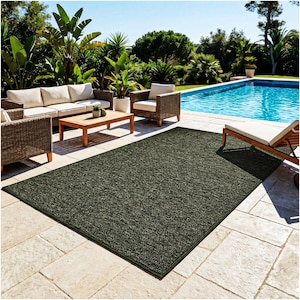 May include: A rectangular area rug with a dark green, textured, low-pile surface and a solid dark green border. The rug is positioned flat on a light-colored stone patio surface, featuring a consistent, dense weave pattern across its entire top.