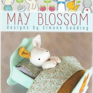May include: A white stuffed bunny rabbit lying in a light blue bed with a green and brown patterned blanket. The bed has a yellow ear on the headboard and the text "rest little rabbit" is printed below the bed.