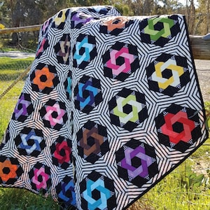 Könnte beinhalten: Eine farbenfrohe Steppdecke mit einem geometrischen Muster aus ineinandergreifenden Sternen. Die Steppdecke ist aus Schwarz, Weiß und verschiedenen leuchtenden Farben gefertigt, darunter Rot, Orange, Gelb, Grün, Blau und Lila.