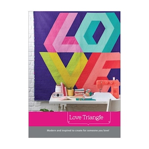 Puede incluir: Un edredón colorido con la palabra "LOVE" en formas geométricas cuelga de una pared morada. Una máquina de coser y un escritorio con suministros están frente al edredón. El texto "Love Triangle" se muestra en la parte inferior.