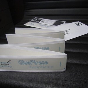 Puede incluir: Un conjunto de tiras adhesivas blancas con el texto "GluePirate Easy Mount" impreso en la tira inferior. Las tiras están dobladas por la mitad y están diseñadas para montar artículos.