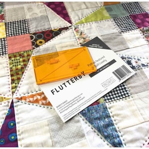 Può includere: Un primo piano di una trapunta con un design a patchwork. La trapunta è realizzata con diversi tessuti di colori e motivi diversi. Un righello da quilting in plastica trasparente arancione è sopra la trapunta. Il righello è etichettato "Flutterby". Un cartoncino bianco con testo nero è parzialmente visibile sullo sfondo. Il cartoncino dice "Modello presentato in Quilt Lovely by Jen Kingwell Designs".