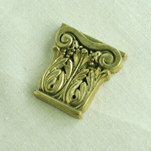 May include: A gold-colored metal decorative piece with an ornate design resembling a column capital. The piece has intricate details of scrolls and leaves.