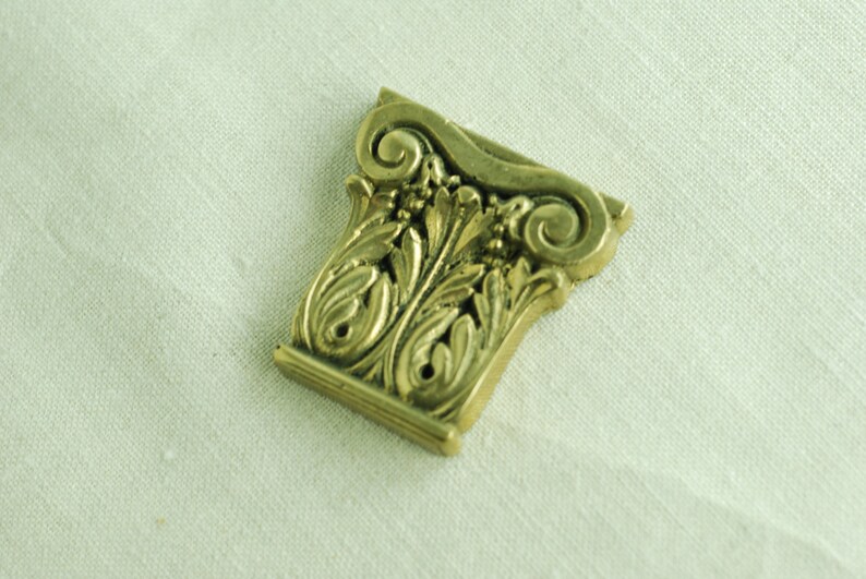 May include: A gold-colored metal decorative piece with an ornate design resembling a column capital. The piece has intricate details of scrolls and leaves.