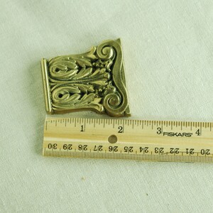 May include: A gold-colored metal decorative piece with an ornate design, measuring approximately 2 inches in length. The piece is shown against a white fabric background.