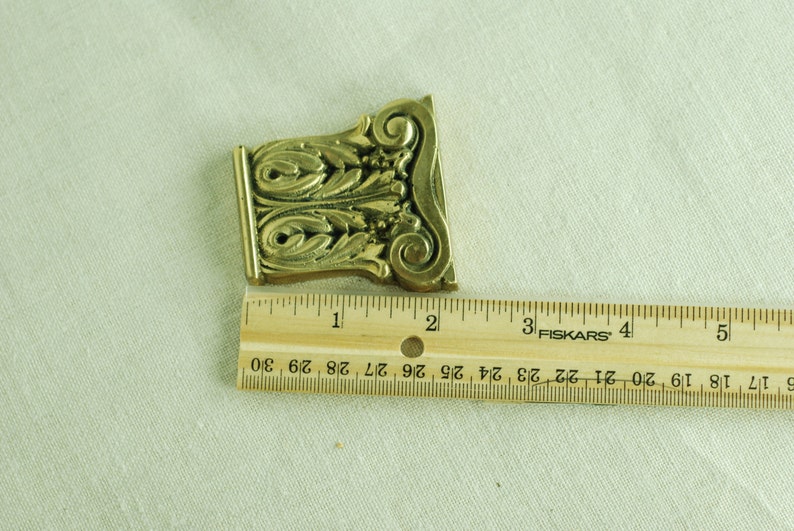 May include: A gold-colored metal decorative piece with an ornate design, measuring approximately 2 inches in length. The piece is shown against a white fabric background.