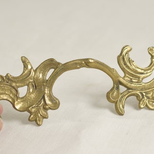 May include: A gold-toned metal decorative hardware piece with a scroll design. The piece has a curved shape and intricate details.