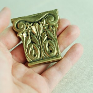 May include: A gold-colored metal decorative piece with intricate leaf and scroll designs. The piece is shaped like a column capital.