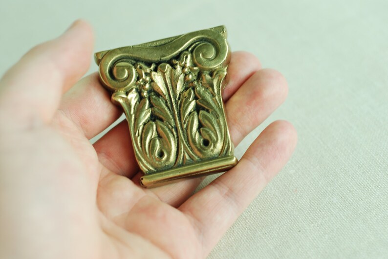 May include: A gold-colored metal decorative piece with intricate leaf and scroll designs. The piece is shaped like a column capital.