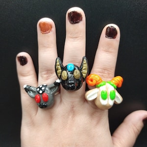 May include: Three polymer clay rings featuring animal faces. The first ring is a black bat with gold accents and a blue gem on its forehead. The second ring is a gray wolf with black spots and red eyes. The third ring is a brown goat with green eyes and orange horns.