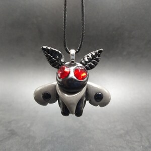 May include: A black and gray moth pendant with red gemstone eyes, hanging from a black cord.