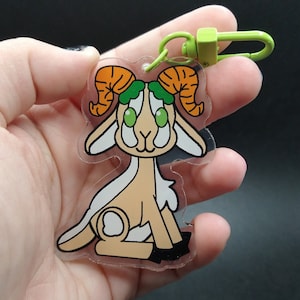 May include: A clear acrylic keychain featuring a cartoon goat with orange horns, green leaves on its head, and large green eyes. The goat is sitting down with its tail curled up.