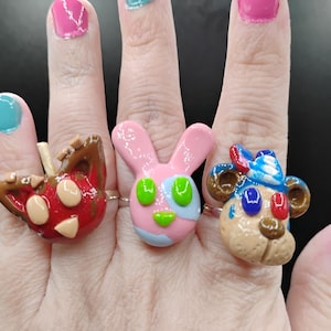 May include: Three handmade rings with cartoon animal faces: a red apple, a pink bunny, and a tan bear. The rings are on a silver band. The bunny has green eyes and blue and pink accents. The bear has a blue and white hat.