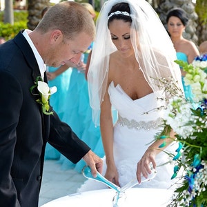 May include: A bride and groom pour blue sand into a glass vase during a wedding ceremony. The bride is wearing a white wedding dress with a veil and the groom is wearing a black suit. The ceremony is taking place outdoors with a floral arrangement in the background.
