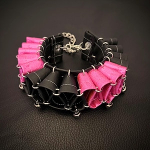 May include: A choker necklace constructed from alternating black and metallic pink folded leather-like segments. The pieces are connected by silver-toned metal jump rings, featuring a black base strap and a silver-toned chain closure at the back.