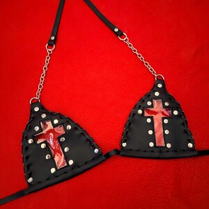 以下が含まれることがあります： 縁にシルバーの金属製スタッドが施された2つの三角形のカップを持つ黒いレザーのビキニトップ。各カップには十字型の切り抜きがあり、その下に赤いマーブル模様の素材が見えます。シルバーのチェーンストラップが付いています。