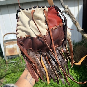 May include: A small, brown leather bag with fringe. The bag is made of different colored leather pieces, including white, brown, and red. The bag has a drawstring closure and a long leather strap.