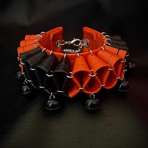 May include: A unique orange and black leather bracelet with a silver clasp. The bracelet features a pleated design and is adorned with small black bells hanging from silver rings. The background is a dark, textured surface.