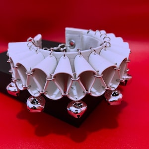 May include: A white collar accessory featuring a series of folded, pleated segments connected by silver-toned metal rings. The lower edge of the collar is adorned with multiple small, round, silver-toned jingle bells hanging from the metal rings.