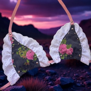 May include: A floral print bikini top with a corduroy texture, featuring pink roses and gold studs. The top has white lace trim and pink straps, set against a purple and pink sunset background.