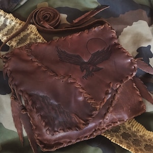 May include: A brown leather crossbody bag with a stitched design. The bag features an eagle design with a sun in the background. The bag has a fringe detail and a long strap.