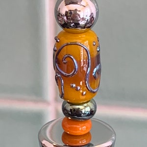 May include: A silver bottle stopper with a decorative top made of glass beads. The top features a round silver bead, a yellow and brown glass bead with a swirl design, a silver bead, and an orange glass bead.