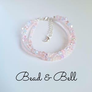 May include: A multi-strand bracelet featuring translucent pink and iridescent beads, accented with a silver-toned metal chain and a small crescent moon charm. The text Bead & Bell is printed in black script below the jewellery piece.