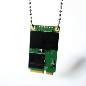 May include: A green circuit board pendant with black components and gold connectors, suspended from a silver ball chain. The pendant has the text "link media" printed on it. The pendant is a unique piece of tech jewelry.