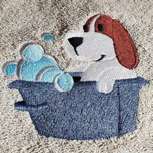 May include: Embroidered beige towel featuring a cartoon dog in a blue tub with blue bubbles. The dog has white fur, brown ears, and a black nose. The towel has a textured appearance.