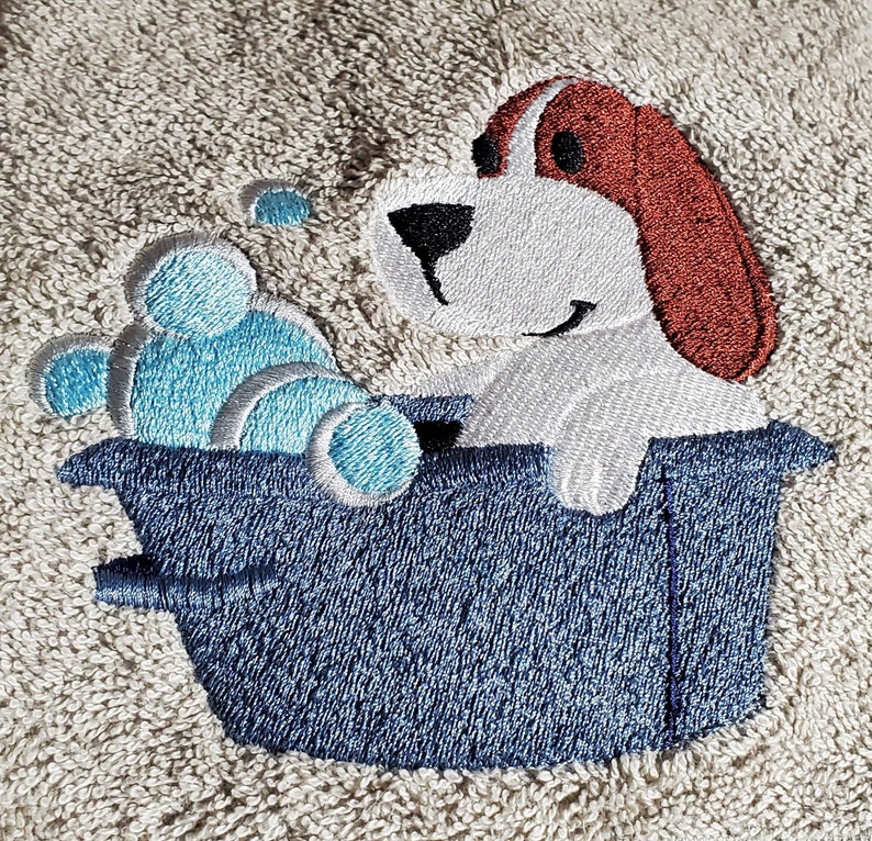 May include: Embroidered beige towel featuring a cartoon dog in a blue tub with blue bubbles. The dog has white fur, brown ears, and a black nose. The towel has a textured appearance.