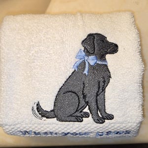 May include: White hand towel with an embroidered black dog wearing a light blue bow. The towel has the phrase "Wash your paws" embroidered in light blue at the bottom. The towel is folded.