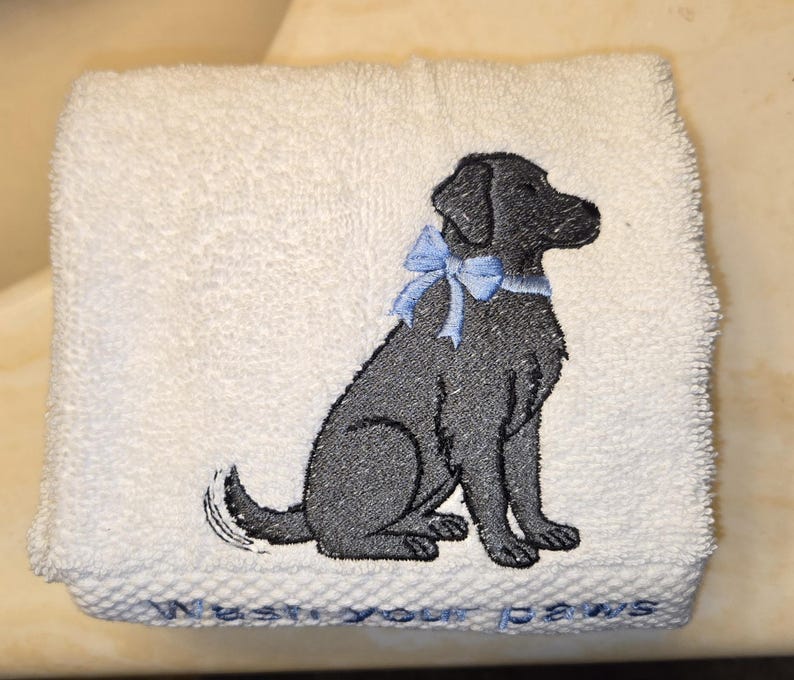 May include: White hand towel with an embroidered black dog wearing a light blue bow. The towel has the phrase "Wash your paws" embroidered in light blue at the bottom. The towel is folded.
