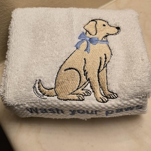 May include: White folded hand towel with an embroidered image of a tan Labrador Retriever wearing a light blue bow. The words "Wash your paws" are embroidered in light blue at the bottom of the towel.