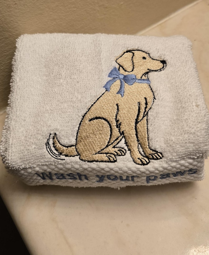 May include: White folded hand towel with an embroidered image of a tan Labrador Retriever wearing a light blue bow. The words "Wash your paws" are embroidered in light blue at the bottom of the towel.