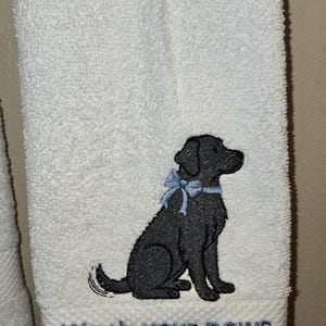 May include: White bath towel with an embroidered black Labrador Retriever wearing a blue bow. The towel has the phrase "Wash your paws" embroidered in blue.