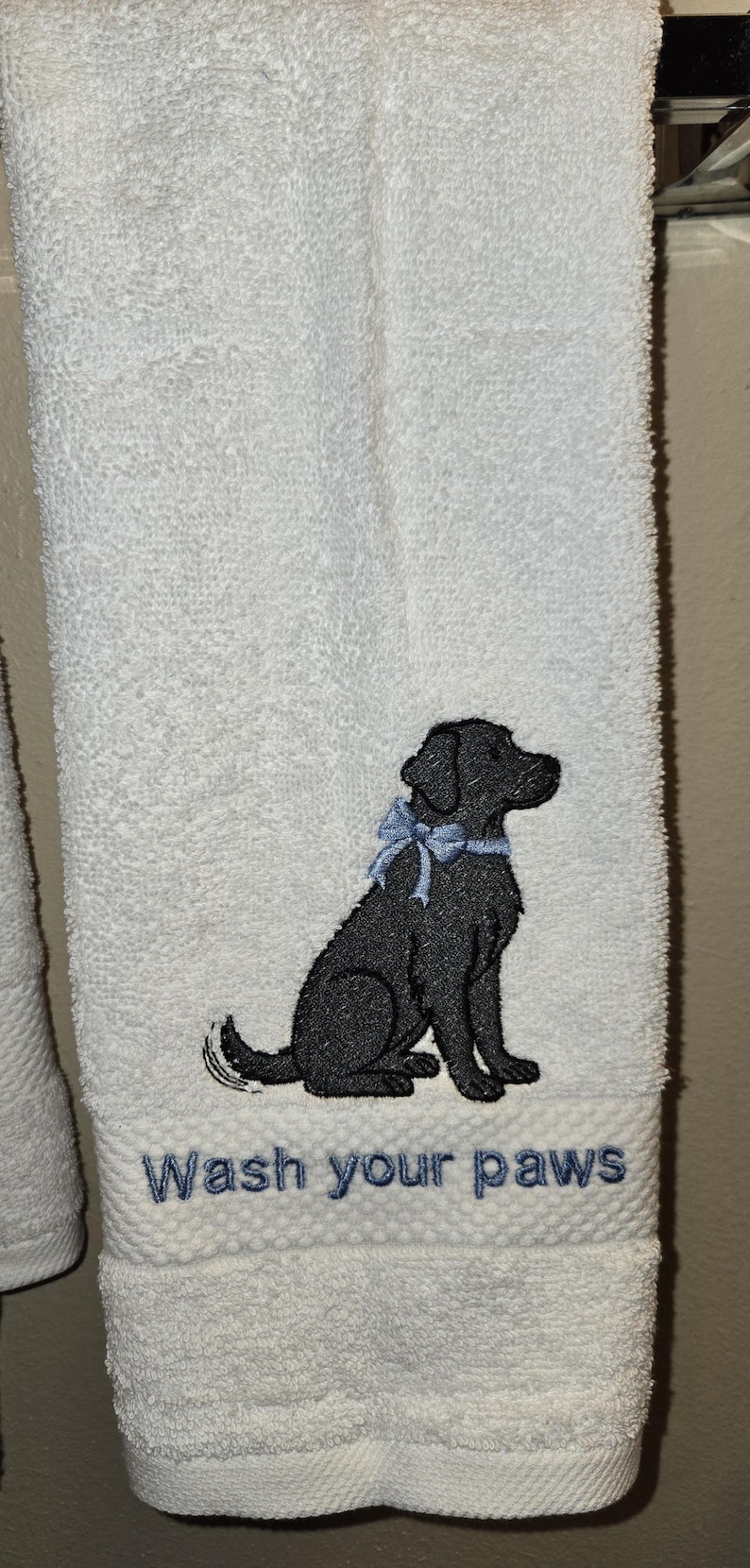 May include: White bath towel with an embroidered black Labrador Retriever wearing a blue bow. The towel has the phrase "Wash your paws" embroidered in blue.