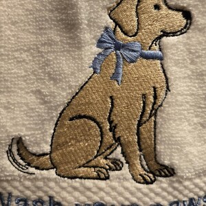 May include: Embroidered white towel featuring a tan dog with a blue bow. The dog is sitting, with the text "Wash your paws" embroidered in blue below. The towel has a textured surface.